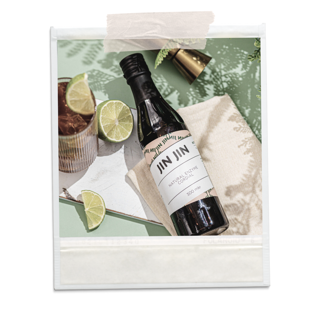 A polaroid of a 300 ml glass bottle of a drink mixer, laying on a picnic blanket alongside a glass and some sliced limes. 